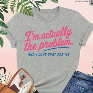 K&ouml;nnte beinhalten: Graues T-Shirt mit dem Text "I'm actually the problem. AND I LOVE THAT FOR ME" in Pink und Blau. Das Shirt wird mit einer blauen Jeans, einer braunen Handtasche und schwarzen High Heels pr&auml;sentiert.