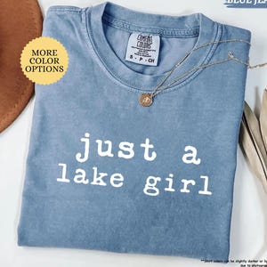 May include: A blue jean colored t-shirt with the text "just a lake girl" in white. The shirt has a crew neck and short sleeves. A gold necklace with a pendant is draped across the shirt. The shirt is labeled "Comfort Colors".