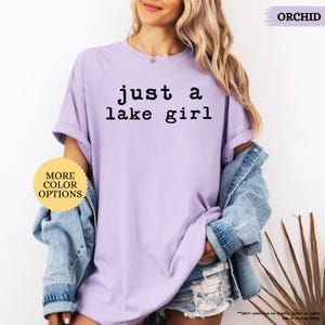 May include: A light purple t-shirt with the text "just a lake girl" in black. The shirt is part of a casual outfit, paired with a denim jacket and shorts. The image also includes a button that says "MORE COLOR OPTIONS".