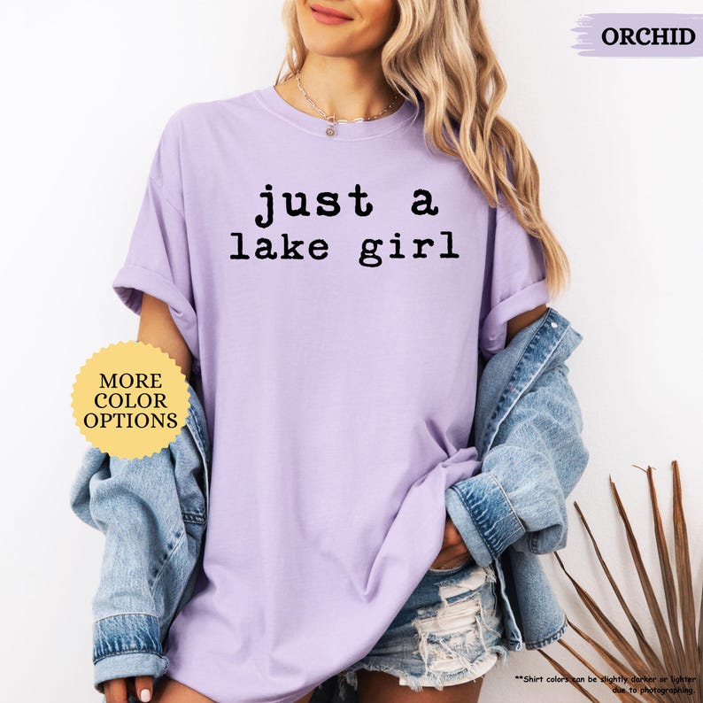 May include: A light purple t-shirt with the text "just a lake girl" in black. The shirt is part of a casual outfit, paired with a denim jacket and shorts. The image also includes a button that says "MORE COLOR OPTIONS".
