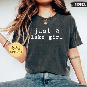 May include: A dark gray t-shirt with the text "just a lake girl" in white. The shirt has a crew neck and short sleeves. A small, round sticker with "MORE COLOR OPTIONS" is visible. The word "PEPPER" is printed on the top right.