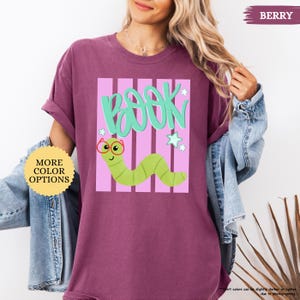May include: A berry-colored t-shirt featuring a graphic of a light green worm wearing red glasses. Above the worm, the word BOOK is written in large teal letters. The design includes light pink vertical stripes and small white star accents.