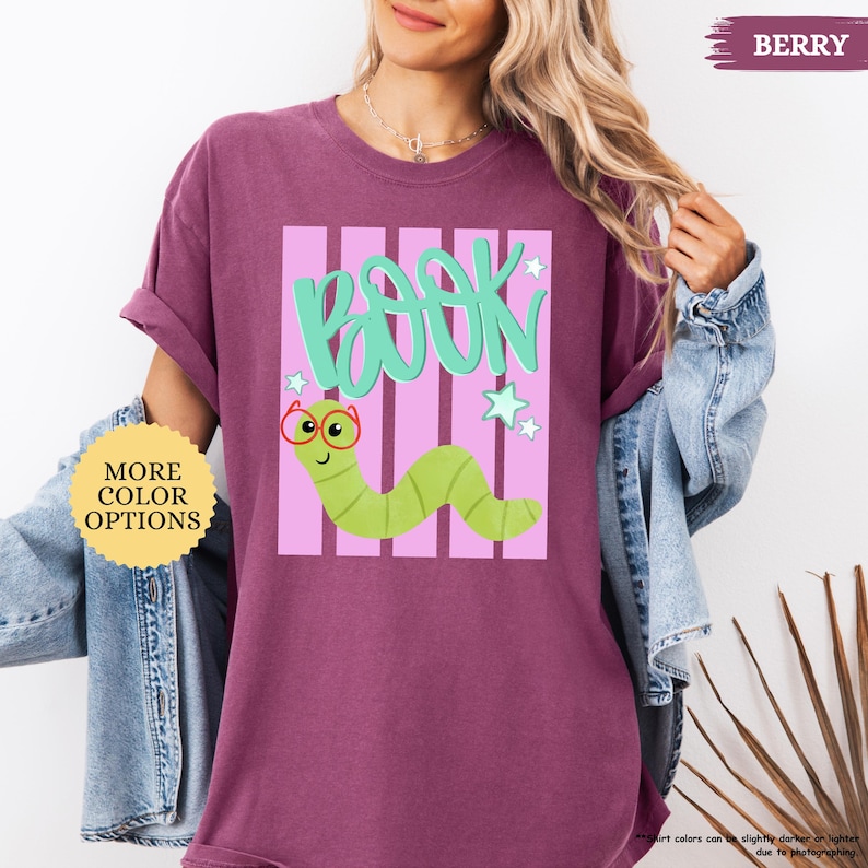 May include: A berry-colored t-shirt featuring a graphic of a light green worm wearing red glasses. Above the worm, the word BOOK is written in large teal letters. The design includes light pink vertical stripes and small white star accents.