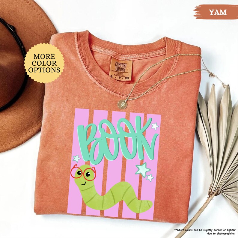 May include: A rust-colored short-sleeve t-shirt featuring a graphic print of a green worm wearing red glasses. The text "BOOK" is written in large teal letters above the worm, set against a background of vertical pink stripes and small white stars.