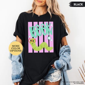 May include: A black t-shirt featuring a graphic design with the word BOOK in large teal letters above a light green cartoon worm wearing red glasses. The design includes vertical pink stripes and small teal stars on a black background.