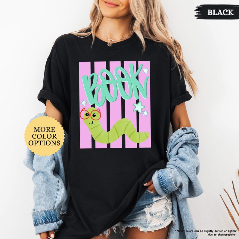 May include: A black t-shirt featuring a graphic design with the word BOOK in large teal letters above a light green cartoon worm wearing red glasses. The design includes vertical pink stripes and small teal stars on a black background.