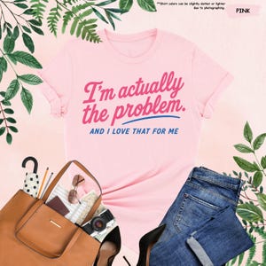 K&ouml;nnte beinhalten: Hellrosa T-Shirt mit dem Text "I'm actually the problem. AND I LOVE THAT FOR ME" in Rosa und Blau. Das Bild zeigt blaue Jeans, eine braune Handtasche, schwarze Abs&auml;tze und Accessoires.
