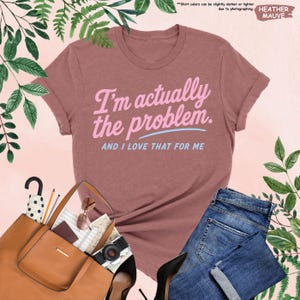 K&ouml;nnte beinhalten: Mauvefarbenes T-Shirt mit dem Text "I'm actually the problem. AND I LOVE THAT FOR ME" in Rosa und Hellblau. Das Shirt ist mit blauen Jeans, einer braunen Handtasche, schwarzen Abs&auml;tzen und Accessoires gestylt.