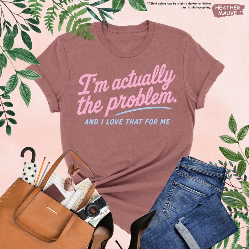 K&ouml;nnte beinhalten: Mauvefarbenes T-Shirt mit dem Text "I'm actually the problem. AND I LOVE THAT FOR ME" in Rosa und Hellblau. Das Shirt ist mit blauen Jeans, einer braunen Handtasche, schwarzen Abs&auml;tzen und Accessoires gestylt.