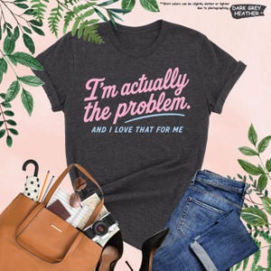 K&ouml;nnte beinhalten: Dunkelgraues Heather-T-Shirt mit dem Text "I'm actually the problem. AND I LOVE THAT FOR ME" in Rosa und Hellblau. Das Shirt wird mit einer blauen Jeans, einer braunen Handtasche und schwarzen Abs&auml;tzen kombiniert.
