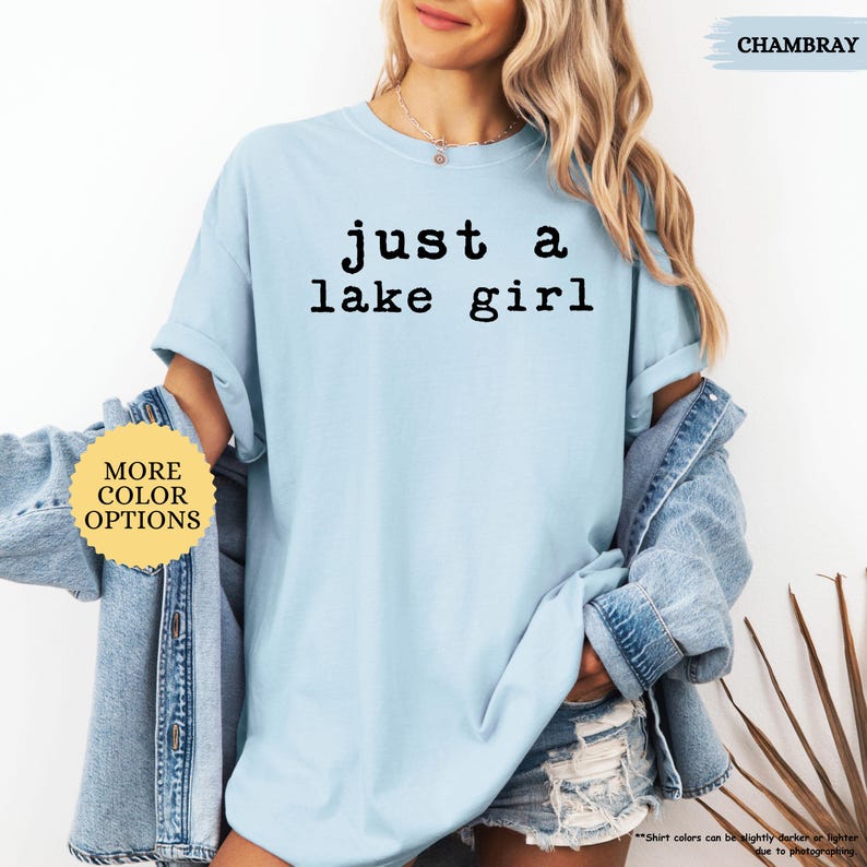 May include: Light blue t-shirt with the text "just a lake girl" in black. The shirt is part of a casual wear collection. The model is wearing a denim jacket and shorts. The words "More Color Options" are visible.