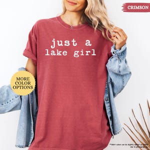 May include: Crimson-colored t-shirt with the text "just a lake girl" in white. The shirt has short sleeves and a relaxed fit. The model is wearing a denim jacket over the shirt. The image also includes a button that says "MORE COLOR OPTIONS".