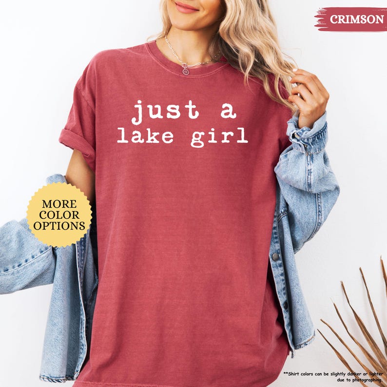 May include: Crimson-colored t-shirt with the text "just a lake girl" in white. The shirt has short sleeves and a relaxed fit. The model is wearing a denim jacket over the shirt. The image also includes a button that says "MORE COLOR OPTIONS".