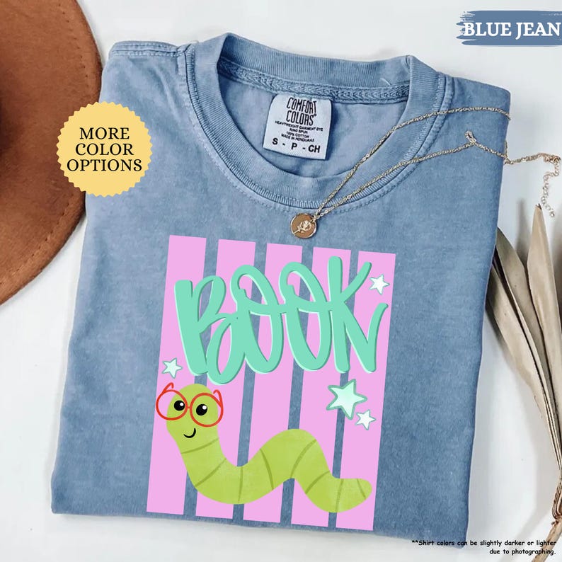 May include: A blue short-sleeve t-shirt featuring a graphic of a green worm wearing red glasses. The design includes the word BOOK in large mint green letters over a background of vertical pink stripes and small white stars.