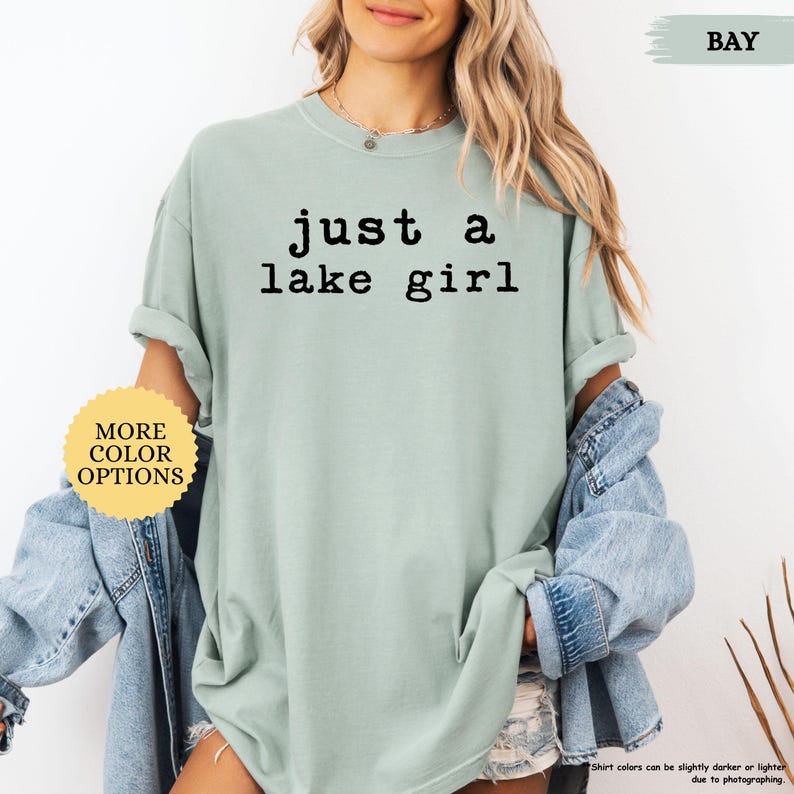 May include: A light green t-shirt with the text "just a lake girl" in black. The shirt has rolled-up sleeves and is paired with a denim jacket. A yellow circle with "MORE COLOR OPTIONS" is in the lower left corner.