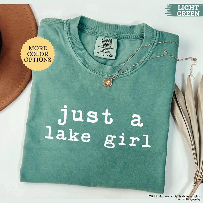 May include: A light green Comfort Colors t-shirt with the text "just a lake girl" in white. A silver necklace with a pendant is draped on the shirt. The shirt is a size small.