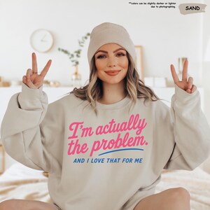 K&ouml;nnte beinhalten: Sandfarbener Pullover mit dem Text "I'm actually the problem. AND I LOVE THAT FOR ME" in Pink und Blau. Das Model tr&auml;gt eine passende M&uuml;tze und zeigt das Victory-Zeichen.