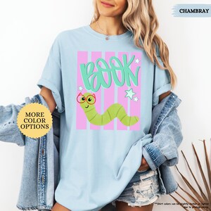 May include: A light blue short-sleeve t-shirt featuring a graphic of a green worm wearing red glasses. Above the worm, the word BOOK is written in a stylized green font. The design includes pink vertical stripes and small white stars.