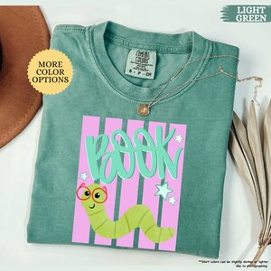 May include: A light green t-shirt featuring a graphic of a green worm wearing red glasses. The text "BOOK" is printed in large light green letters above the worm. The design includes pink vertical stripes and small white stars on the shirt.