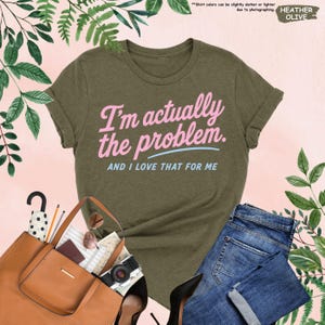 K&ouml;nnte beinhalten: Olivgr&uuml;nes T-Shirt mit dem Text "I'm actually the problem. AND I LOVE THAT FOR ME" in Rosa und Blau. Das Shirt wird mit einer blauen Jeans, einer braunen Tragetasche, schwarzen Abs&auml;tzen und Accessoires kombiniert. Das Shirt ist aus weichem Material.