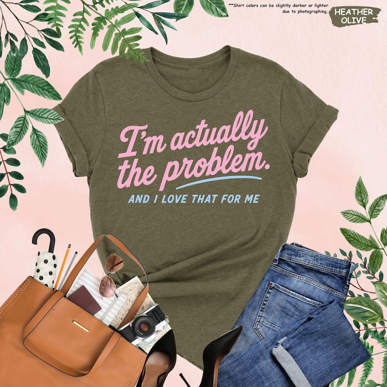 K&ouml;nnte beinhalten: Olivgr&uuml;nes T-Shirt mit dem Text "I'm actually the problem. AND I LOVE THAT FOR ME" in Rosa und Blau. Das Shirt wird mit einer blauen Jeans, einer braunen Tragetasche, schwarzen Abs&auml;tzen und Accessoires kombiniert. Das Shirt ist aus weichem Material.