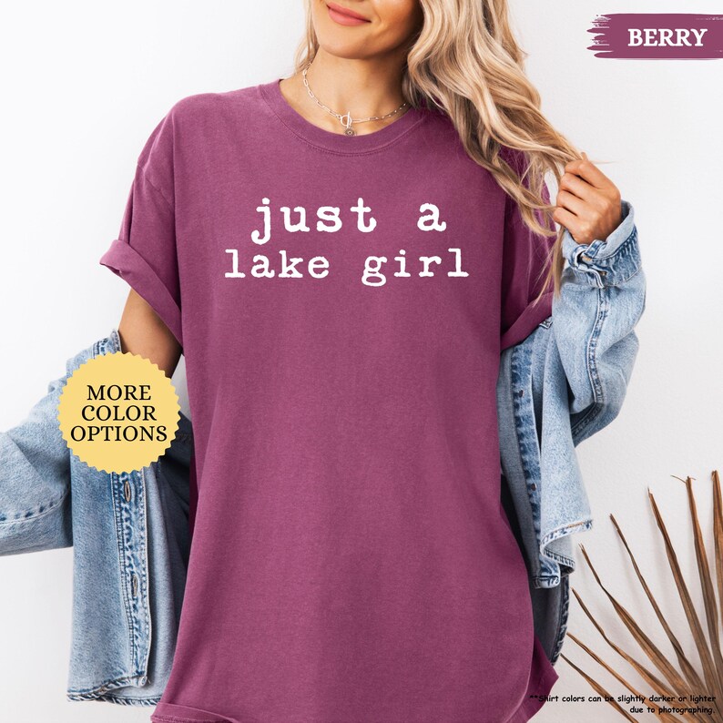 May include: Berry-colored t-shirt with the text "just a lake girl" in white. The shirt has rolled-up sleeves and a round neckline. A denim jacket is worn over the shirt. A button on the shirt says "MORE COLOR OPTIONS".