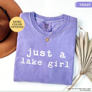May include: A violet Comfort Colors t-shirt with the text "just a lake girl" in white. A gold necklace with a pendant rests on the shirt. The shirt is displayed with a brown hat and a dried plant.