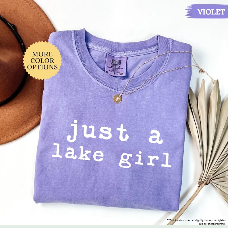 May include: A violet Comfort Colors t-shirt with the text "just a lake girl" in white. A gold necklace with a pendant rests on the shirt. The shirt is displayed with a brown hat and a dried plant.