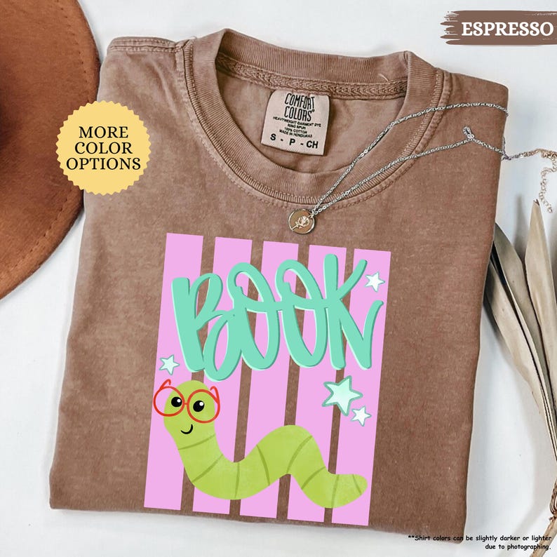 May include: An espresso-colored t-shirt featuring a graphic of a green worm wearing red glasses. The word BOOK is written in large teal script above the worm, set against a background of vertical pink stripes and small white stars.
