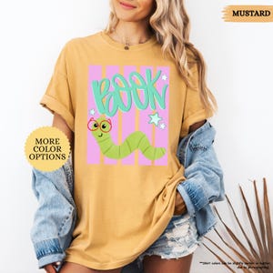 May include: A mustard yellow t-shirt featuring a graphic print of a green worm wearing red glasses. The design includes the word BOOK in large teal letters above the worm, set against a background of vertical pink stripes and small white stars.