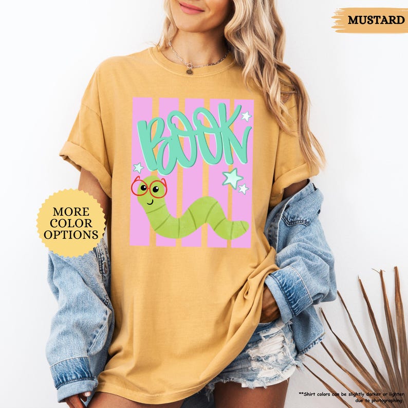 May include: A mustard yellow t-shirt featuring a graphic print of a green worm wearing red glasses. The design includes the word BOOK in large teal letters above the worm, set against a background of vertical pink stripes and small white stars.