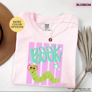 May include: A light pink t-shirt featuring a graphic of a green worm wearing red glasses. The text "BOOK" is printed in large teal letters above the worm. The design includes light pink vertical stripes and small teal stars on a light pink background.