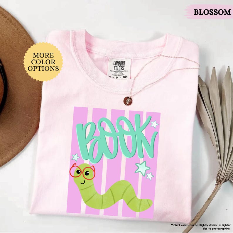 May include: A light pink t-shirt featuring a graphic of a green worm wearing red glasses. The text "BOOK" is printed in large teal letters above the worm. The design includes light pink vertical stripes and small teal stars on a light pink background.