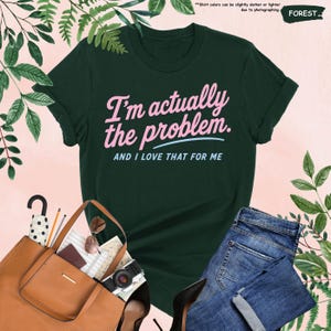 K&ouml;nnte beinhalten: Forstgr&uuml;nes T-Shirt mit dem Text "I'm actually the problem. AND I LOVE THAT FOR ME" in Rosa und Hellblau. Das Shirt wird mit einer blauen Jeans, einer braunen Handtasche, einer Sonnenbrille und einer Kamera kombiniert.