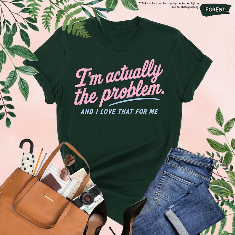 K&ouml;nnte beinhalten: Forstgr&uuml;nes T-Shirt mit dem Text "I'm actually the problem. AND I LOVE THAT FOR ME" in Rosa und Hellblau. Das Shirt wird mit einer blauen Jeans, einer braunen Handtasche, einer Sonnenbrille und einer Kamera kombiniert.