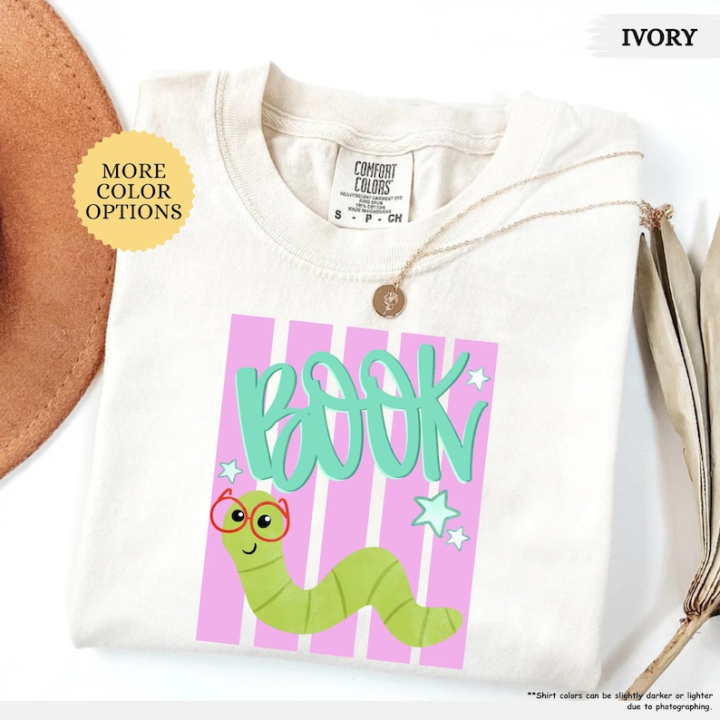 May include: An ivory t-shirt featuring a graphic of a green worm wearing red glasses. Above the worm, the word BOOK is written in large teal letters. The design includes a background of vertical pink stripes and small teal stars.
