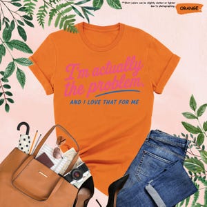 K&ouml;nnte beinhalten: Orangefarbenes T-Shirt mit umgekrempelten &Auml;rmeln und dem Text "I'm actually the problem AND I LOVE THAT FOR ME" in Pink und Blau. Das Bild zeigt blaue Jeans, eine braune Handtasche und weiteres Zubeh&ouml;r.