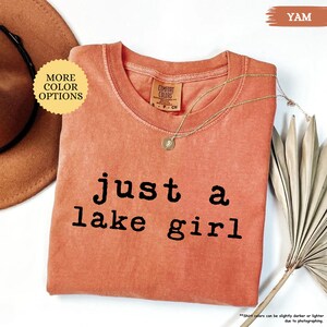 May include: A yam-colored t-shirt with the text "just a lake girl" in black. The shirt has a crew neck and a small tag that says "Comfort Colors". A gold necklace with a pendant is draped across the shirt.