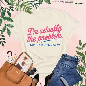 K&ouml;nnte beinhalten: Cremefarbenes T-Shirt mit dem Text "I'm actually the problem. AND I LOVE THAT FOR ME" in Pink und Blau. Das Shirt wird mit blauen Jeans, einer braunen Handtasche, schwarzen Abs&auml;tzen und Accessoires kombiniert. Das Bild hat eine nat&uuml;rliche &Auml;sthetik.