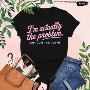 K&ouml;nnte beinhalten: Schwarzes T-Shirt mit dem Text "I'm actually the problem. AND I LOVE THAT FOR ME" in Rosa und Blau. Das Shirt wird mit einer braunen Tragetasche, Sonnenbrille, einer Kamera und einer blauen Jeans pr&auml;sentiert.