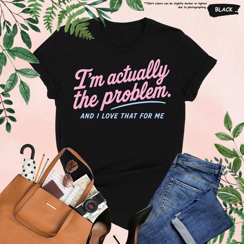 K&ouml;nnte beinhalten: Schwarzes T-Shirt mit dem Text "I'm actually the problem. AND I LOVE THAT FOR ME" in Rosa und Blau. Das Shirt wird mit einer braunen Tragetasche, Sonnenbrille, einer Kamera und einer blauen Jeans pr&auml;sentiert.