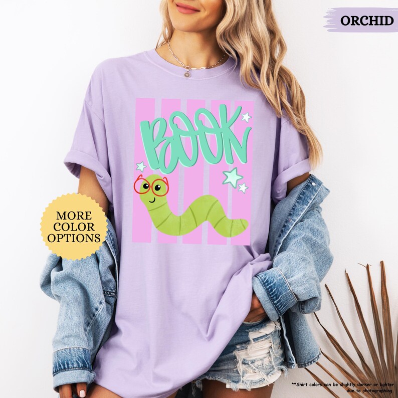 May include: An orchid-colored t-shirt featuring a graphic of a green worm wearing red glasses, the word BOOK in teal, and light teal stars. The design is set against a background of vertical pink stripes. The shirt has short sleeves and a crew neck.