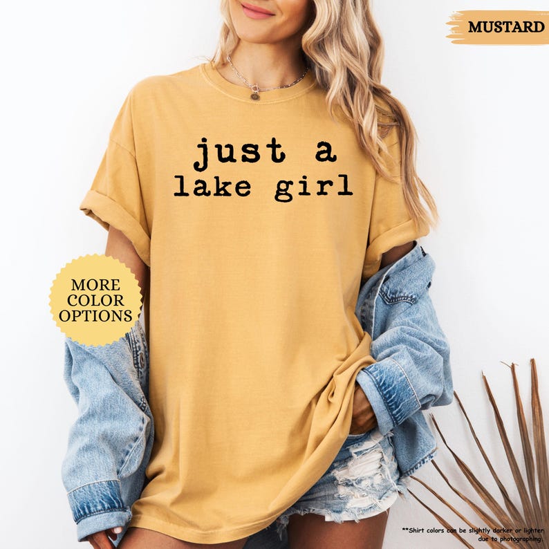 May include: Mustard-colored t-shirt with the text "just a lake girl" in black. The shirt has rolled-up sleeves and is paired with a denim jacket and shorts. A button indicates "More Color Options".