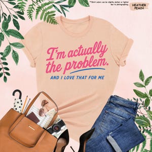 K&ouml;nnte beinhalten: Pfirsichfarbenes T-Shirt mit dem Text "I'm actually the problem. AND I LOVE THAT FOR ME" in Pink und Blau. Das Bild zeigt eine braune Handtasche, Sonnenbrille, eine Kamera und blaue Jeans.