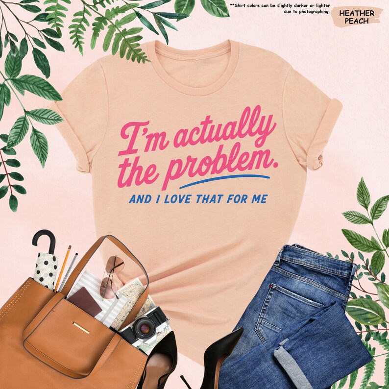 K&ouml;nnte beinhalten: Pfirsichfarbenes T-Shirt mit dem Text "I'm actually the problem. AND I LOVE THAT FOR ME" in Pink und Blau. Das Bild zeigt eine braune Handtasche, Sonnenbrille, eine Kamera und blaue Jeans.