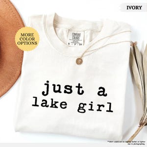 May include: Ivory-colored t-shirt with the text "just a lake girl" in black. The shirt has a crew neck and short sleeves. A gold necklace with a pendant is draped across the shirt. The words "More Color Options" are in a yellow circle.