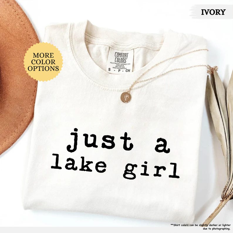 May include: Ivory-colored t-shirt with the text "just a lake girl" in black. The shirt has a crew neck and short sleeves. A gold necklace with a pendant is draped across the shirt. The words "More Color Options" are in a yellow circle.