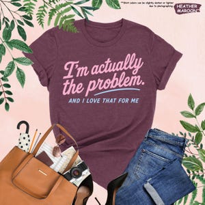 K&ouml;nnte beinhalten: Ein weinrotes T-Shirt mit dem Text "I'm actually the problem. AND I LOVE THAT FOR ME" in Rosa und Hellblau. Das Shirt wird mit einer blauen Jeans, einer braunen Handtasche und schwarzen Abs&auml;tzen kombiniert. Das Wort "HEATHER MAROON" steht oben rechts.