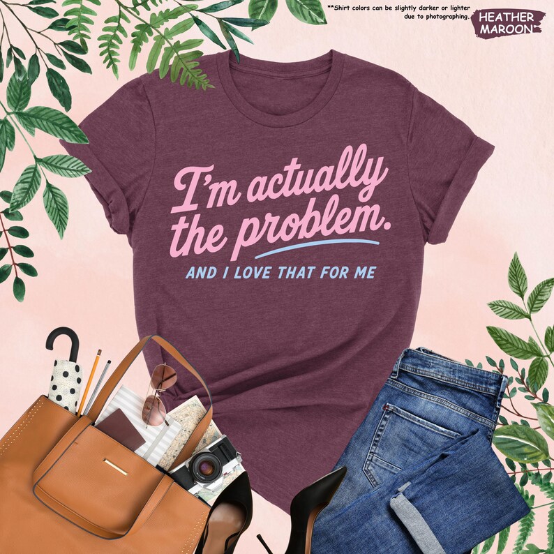 K&ouml;nnte beinhalten: Ein weinrotes T-Shirt mit dem Text "I'm actually the problem. AND I LOVE THAT FOR ME" in Rosa und Hellblau. Das Shirt wird mit einer blauen Jeans, einer braunen Handtasche und schwarzen Abs&auml;tzen kombiniert. Das Wort "HEATHER MAROON" steht oben rechts.