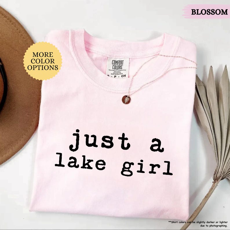 May include: A light pink t-shirt with the text "just a lake girl" in black. The shirt has a crew neck and short sleeves. A gold necklace with a small pendant is draped on the shirt. The word "BLOSSOM" is in the top right corner.